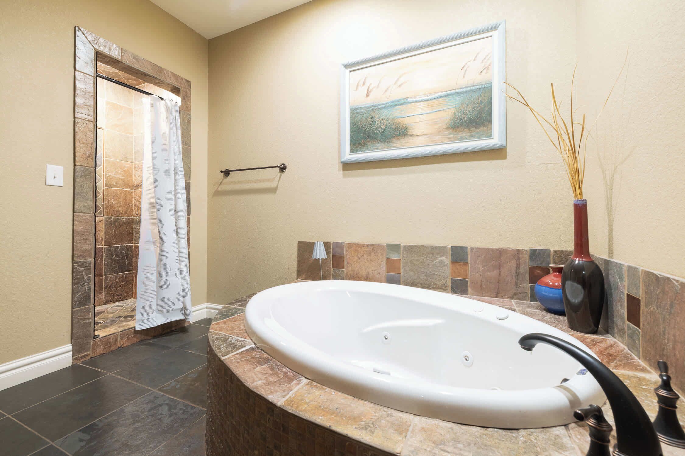 A bathroom with a stone shower enclosure, a bathtub, a towel rack, a framed painting, and a vase with reeds.