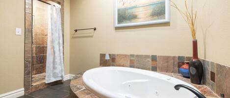 A bathroom with a stone shower enclosure, a bathtub, a towel rack, a framed painting, and a vase with reeds.