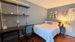 Une chambre à coucher avec un lit, un bureau et une chaise, des étagères, une horloge murale, un attrape-rêves et une porte.
