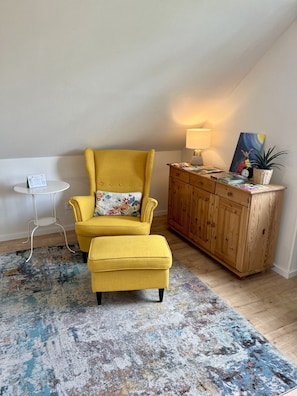 A yellow armchair with a floral pillow, a small white table, a wooden cabinet, and a patterned rug in a room with a sloped ceiling. - Ferienwohnung Wildblume Meißner - Erholung im Meißnervorland (Meißner)