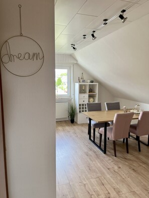 A dining area with a wooden table and chairs, a white cabinet with shelves, and a ceiling track lighting system. - Ferienwohnung Wildblume Meißner - Erholung im Meißnervorland (Meißner)