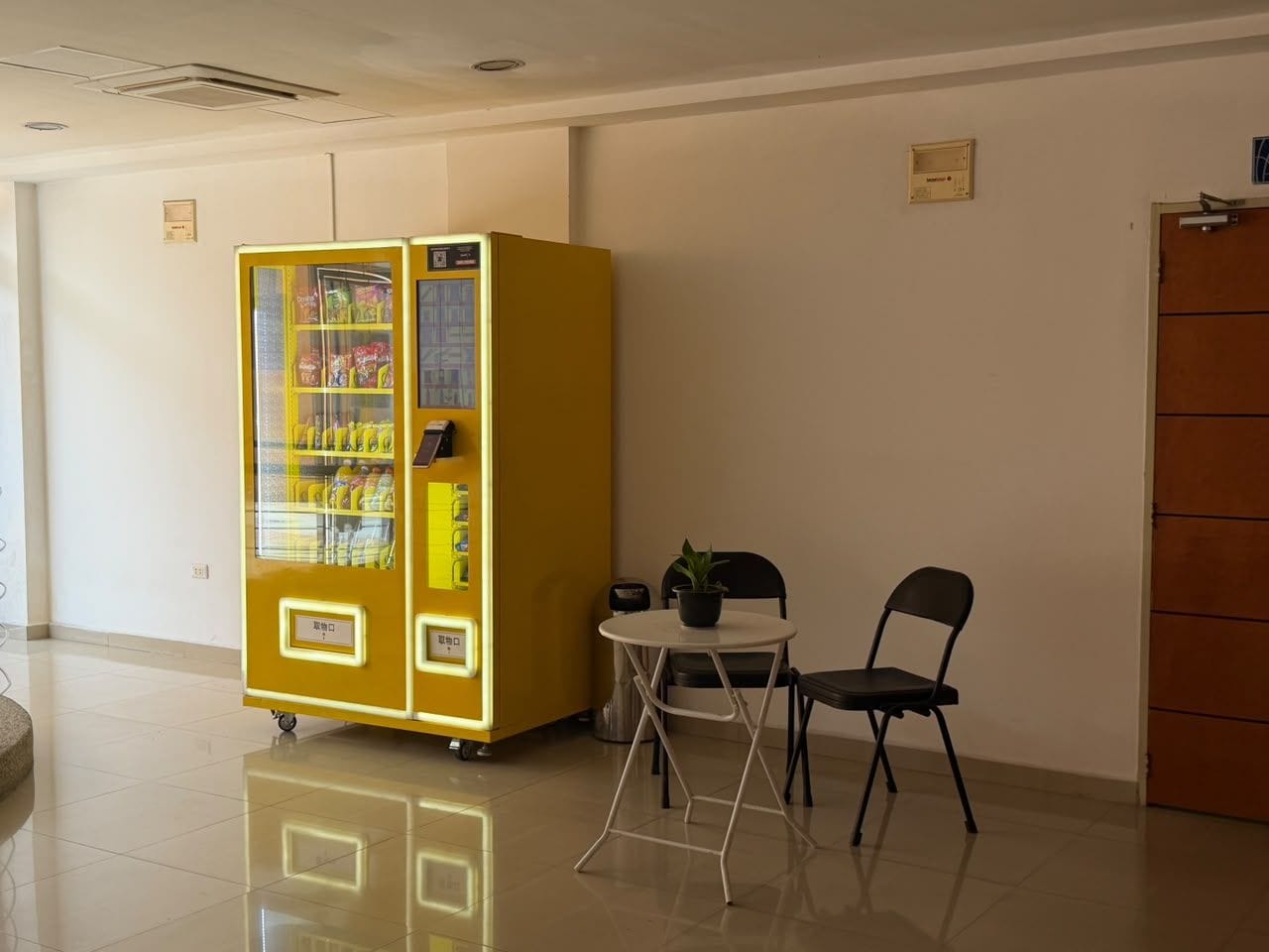 A vending machine, a small table with a chair, and a potted plant in a clean, well-lit indoor space.