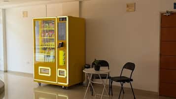 A vending machine, a small table with a chair, and a potted plant in a clean, well-lit indoor space.