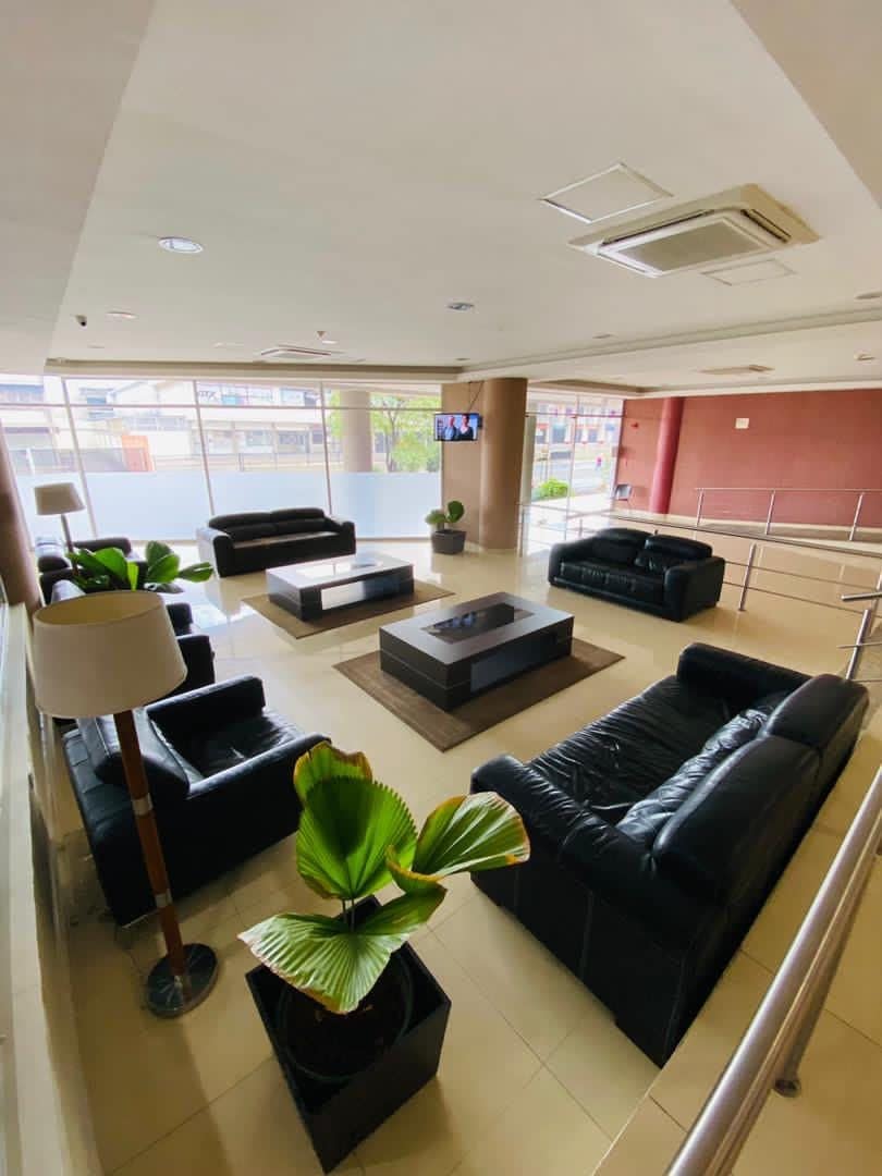 Um hall amplo com sofás de couro preto, uma mesa de centro e plantas em vasos.