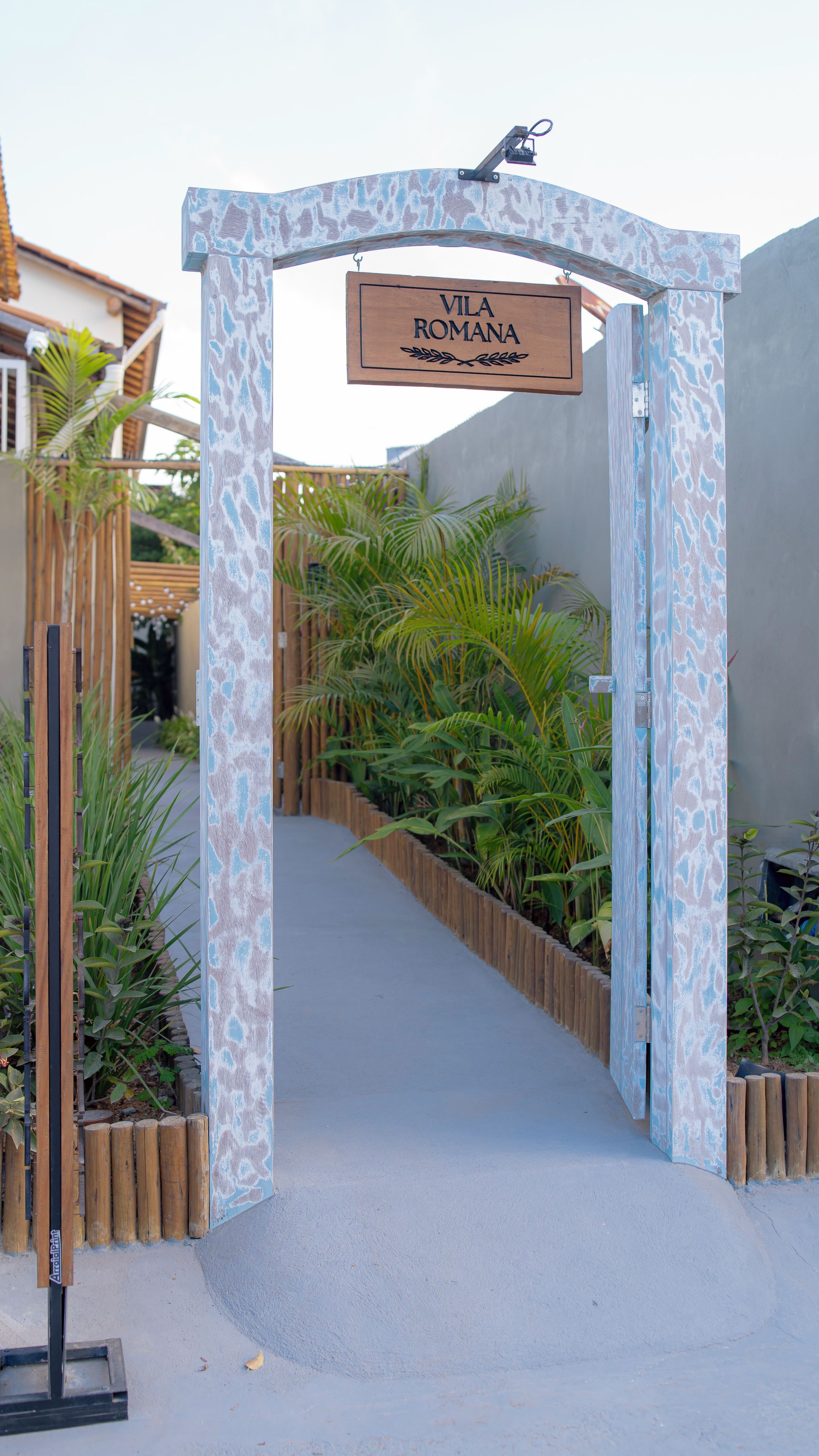 A wooden archway with a sign reading 'Vila Romana' leading to a pathway flanked by wooden fences and green plants.
