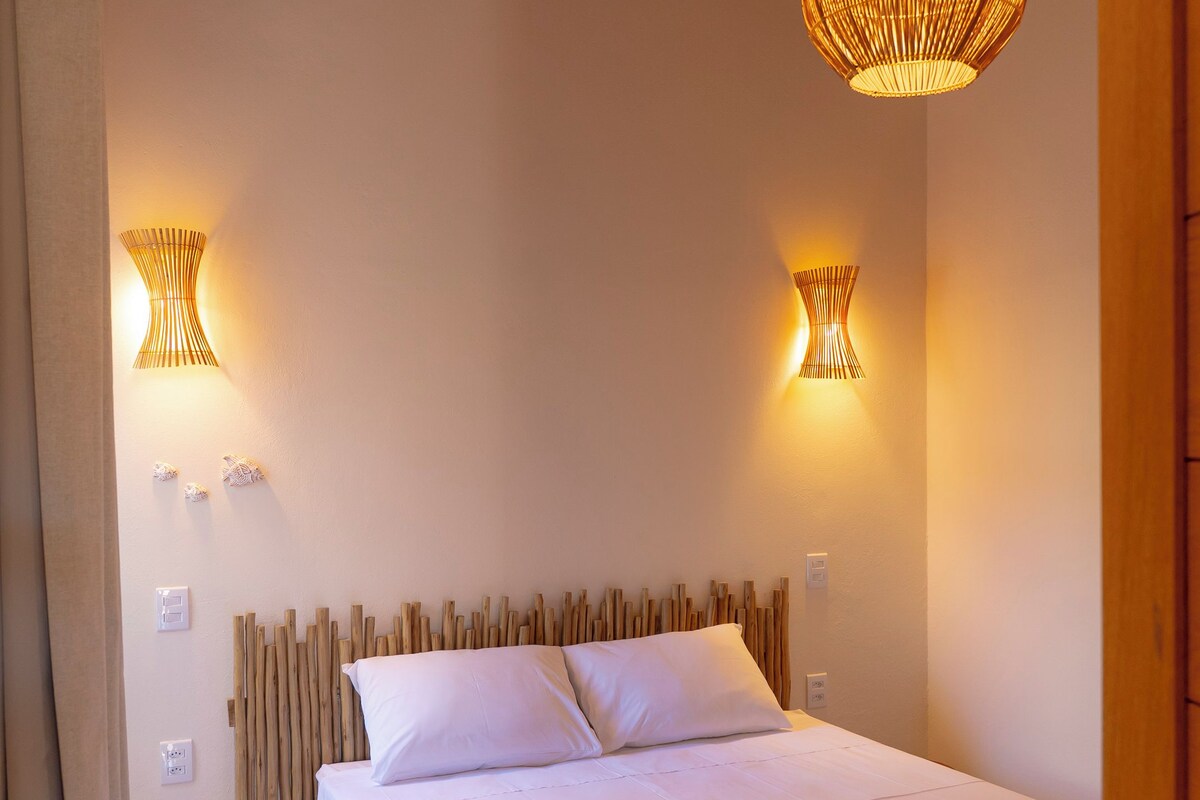 Quarto com cabeceira de madeira, cama com roupa de cama branca, duas luminárias fixadas na parede e porta de madeira.