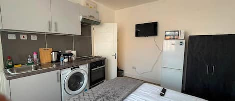 A compact hotel room with a kitchenette, a washing machine, a bed, a TV, and a wardrobe.
