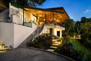 A modern two-story house with a staircase, outdoor lighting, and lush greenery. - Panoramic Ocean Views • Eco-Luxury Solar Retreat with 5 bedrooms (Santa Teresa)