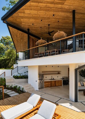 A modern outdoor living area with a wooden deck, lounge chairs, a barbecue grill, and a kitchen area. - Panoramic Ocean Views • Eco-Luxury Solar Retreat with 5 bedrooms (Santa Teresa)