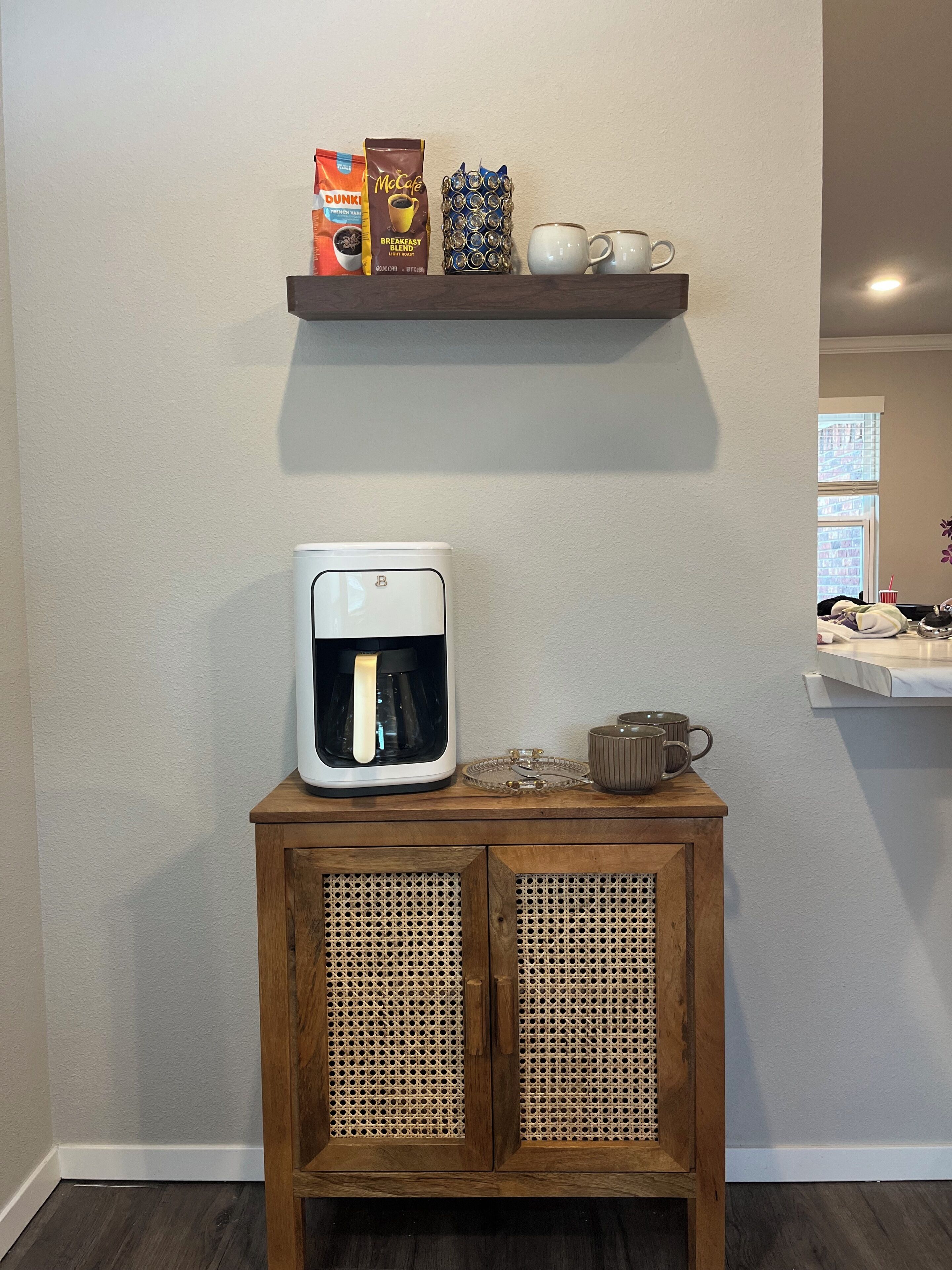 A coffee maker on a wooden cabinet with a shelf above containing a coffee bag, a mug, and a decorative item.