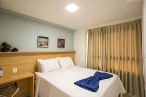 A hotel room with a bed, a headboard, a bedside table, a wall-mounted control panel, two framed pictures, a window with curtains, and a potted plant. - Unigreen Residence Manaira by Carpediem Close to the Beach, Manaira Shopping and MAG Shopping (João Pessoa)
