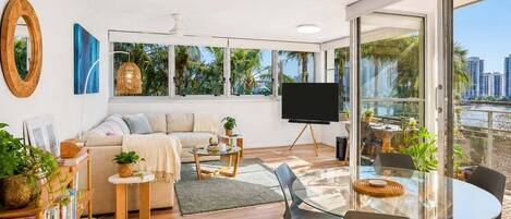 A modern living room with a sofa, coffee table, and chairs, a TV, and large windows with a view of palm trees and buildings.