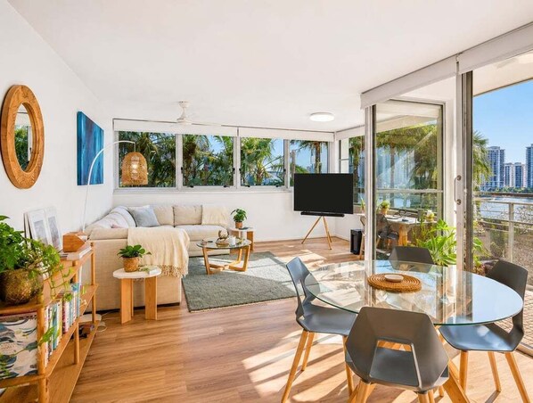 A modern living room with a sofa, coffee table, and chairs, a TV, and large windows with a view of palm trees and buildings. - 2BR Riverside Retreat with Pool Near Beach (Gold Coast)