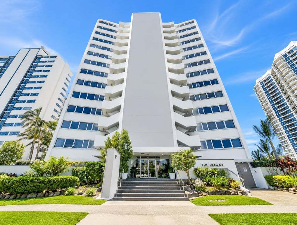 A modern high-rise hotel with a central entrance, multiple balconies, and a well-maintained garden area.