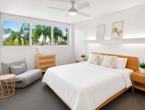A bedroom with a bed, a chair, a nightstand, a ceiling fan, and a window with a view of palm trees. - 2BR Riverside Retreat with Pool Near Beach (Gold Coast)