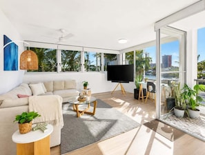 A modern living room with a sofa, coffee table, TV, and large windows. - 2BR Riverside Retreat with Pool Near Beach (Gold Coast)