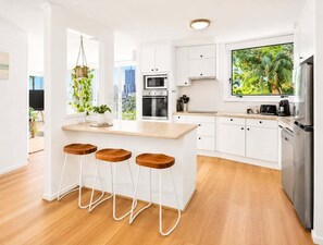 A modern kitchen with white cabinets, a wooden island with stools, a built-in oven, and a large window with a view of greenery. - 2BR Riverside Retreat with Pool Near Beach (Gold Coast)