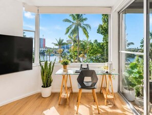 A modern room with a glass table, two chairs, a flat-screen TV, and a large window with a view of palm trees and buildings. - 2BR Riverside Retreat with Pool Near Beach (Gold Coast)