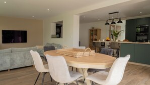 A modern living room with a round dining table, white chairs, a grey sofa, and a kitchen area with dark cabinets and a built-in oven. - Stunning, Renovated Family Villa in Le Pouliguen (Le Pouliguen)