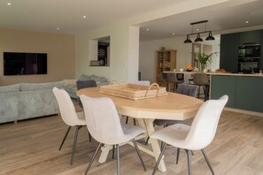 A modern living room with a round dining table, white chairs, a grey sofa, and a kitchen area with dark cabinets and a built-in oven. - Stunning, Renovated Family Villa in Le Pouliguen (Le Pouliguen)