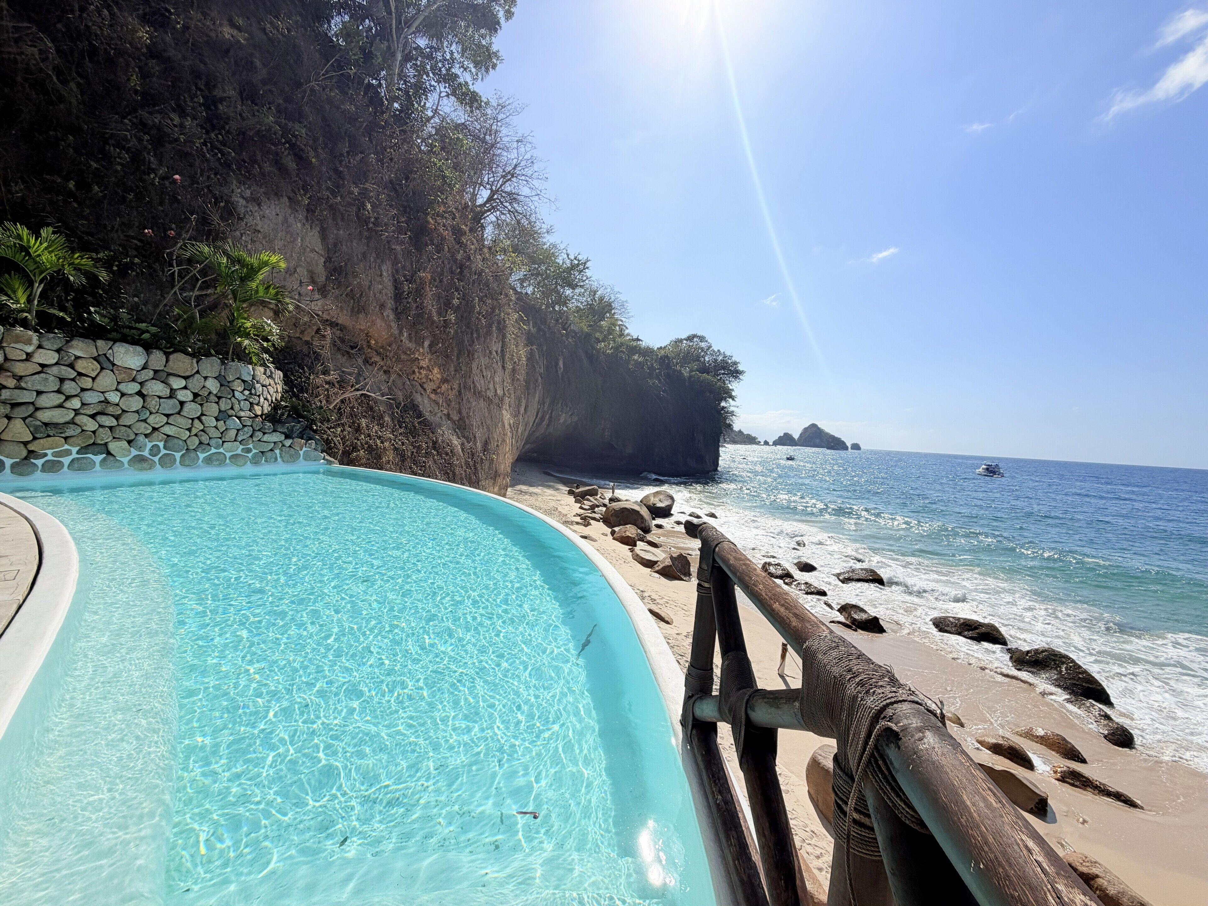 A clear pool with a stone wall, a wooden deck, and a view of the ocean.