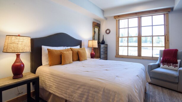 A bedroom with a large bed, two bedside tables with lamps, a dresser, a window with a view, and a chair with a blanket. - Modern Elkhorn Updated 2 bedroom/2 bath (Sun Valley)