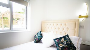 Ein Bett mit einer gebügelten Kopfteile, zwei Kissen mit floralem Muster, ein Nachttisch mit Lampe und ein Fenster mit Blick auf eine Holzpergola.