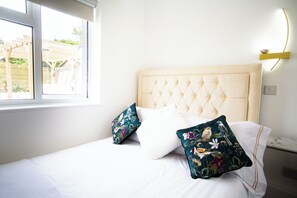 Ein Bett mit einer gebügelten Kopfteile, zwei Kissen mit floralem Muster, ein Nachttisch mit Lampe und ein Fenster mit Blick auf eine Holzpergola.