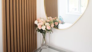 Ein Spiegel mit goldener Rahmung, eine runde Leuchte darüber und eine Vase mit Blumen auf einem Glastisch.