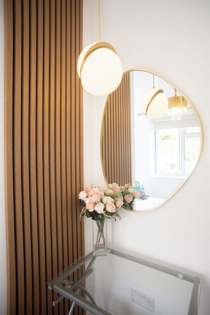 Ein Spiegel mit goldener Rahmung, eine runde Leuchte darüber und eine Vase mit Blumen auf einem Glastisch.