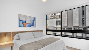 Ett modernt hotellrum med en säng, ett nattduksbord, ett fönster med utsikt över byggnader och ett abstrakt konstverk på väggen.