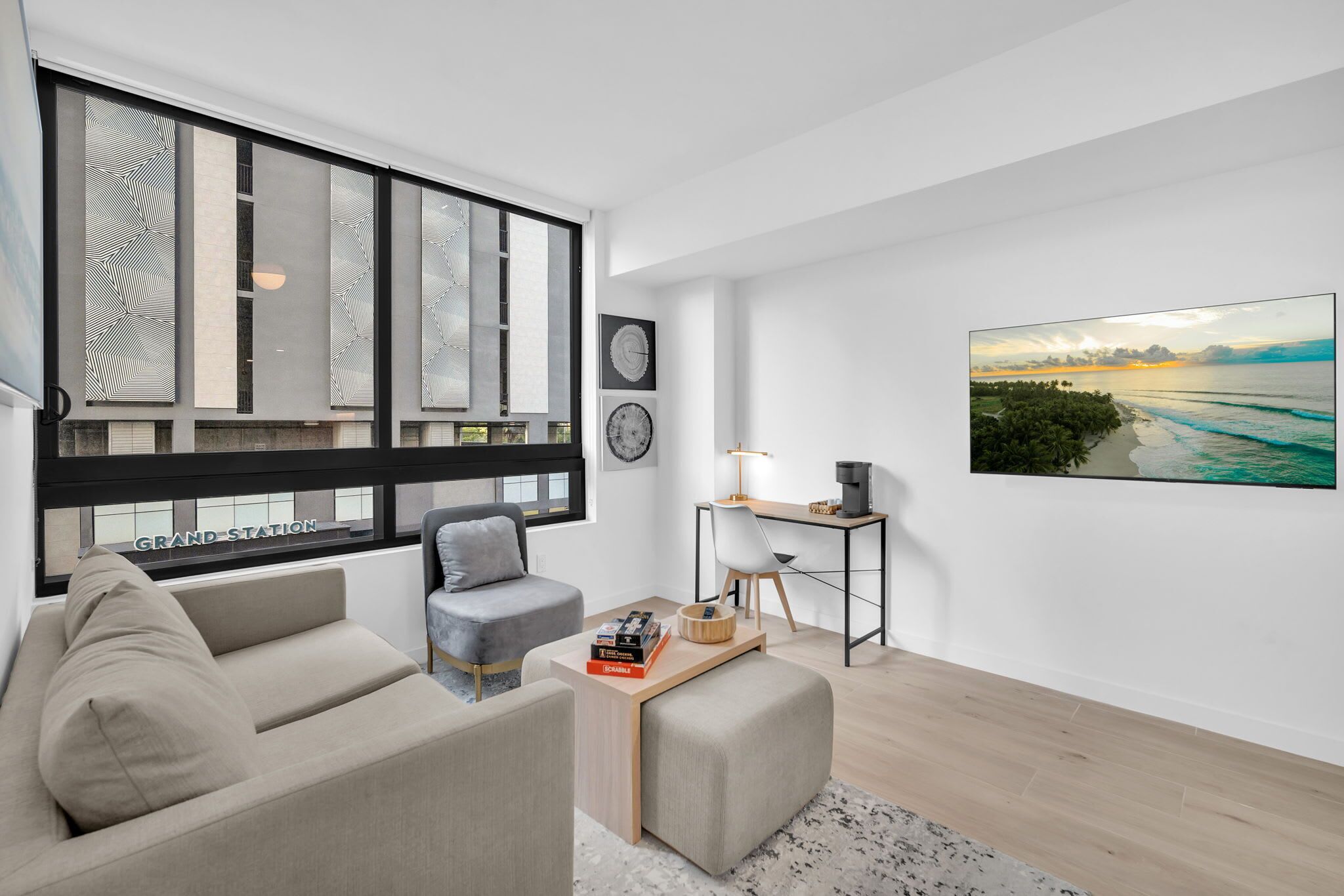 Ein modernes Wohnzimmer mit einer Couch, einem Couchtisch und einem Schreibtisch mit Lampe. Große Fenster bieten einen Blick ins Freie.
