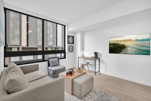 A modern living room with a sofa, a coffee table, and a desk with a lamp. There are large windows and a view of the outside. - Dazzling 2BR Miami | Rooftop Pool & Co-Working (Miami)