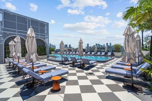 Ein Poolbereich auf dem Dach mit Liegestühlen und Sonnenschirmen, einem Schachbrettmuster auf dem Boden und einer Stadtansicht im Hintergrund.