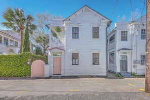 Une maison blanche à deux étages, avec une porte rose, un porche couvert et un petit jardin devant, bordé d’une haie.