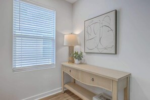 Ein Zimmer mit Fenster, Jalousien, einer Lampe auf einer Konsole und einem gerahmten abstrakten Kunstwerk an der Wand.