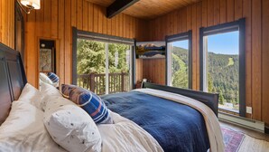 Ein Schlafzimmer mit Holzvertäfelung, einem Bett, Kissen und Blick auf einen Wald durch große Fenster.