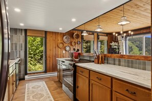 A kitchen with wooden cabinets, a stove, and a large window with a view of greenery. - Schweitzer Mtn. Retreat -Chalet 75-w/Hot Tub Room + Lift Pass (Sandpoint)