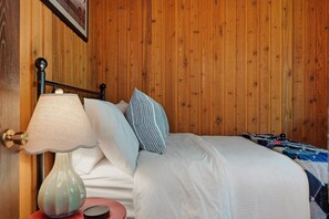 Een wand van houten panelen, een bed met witte beddengoed en gestreepte kussens, een lamp en een klein rode tafel met een roze voorwerp.