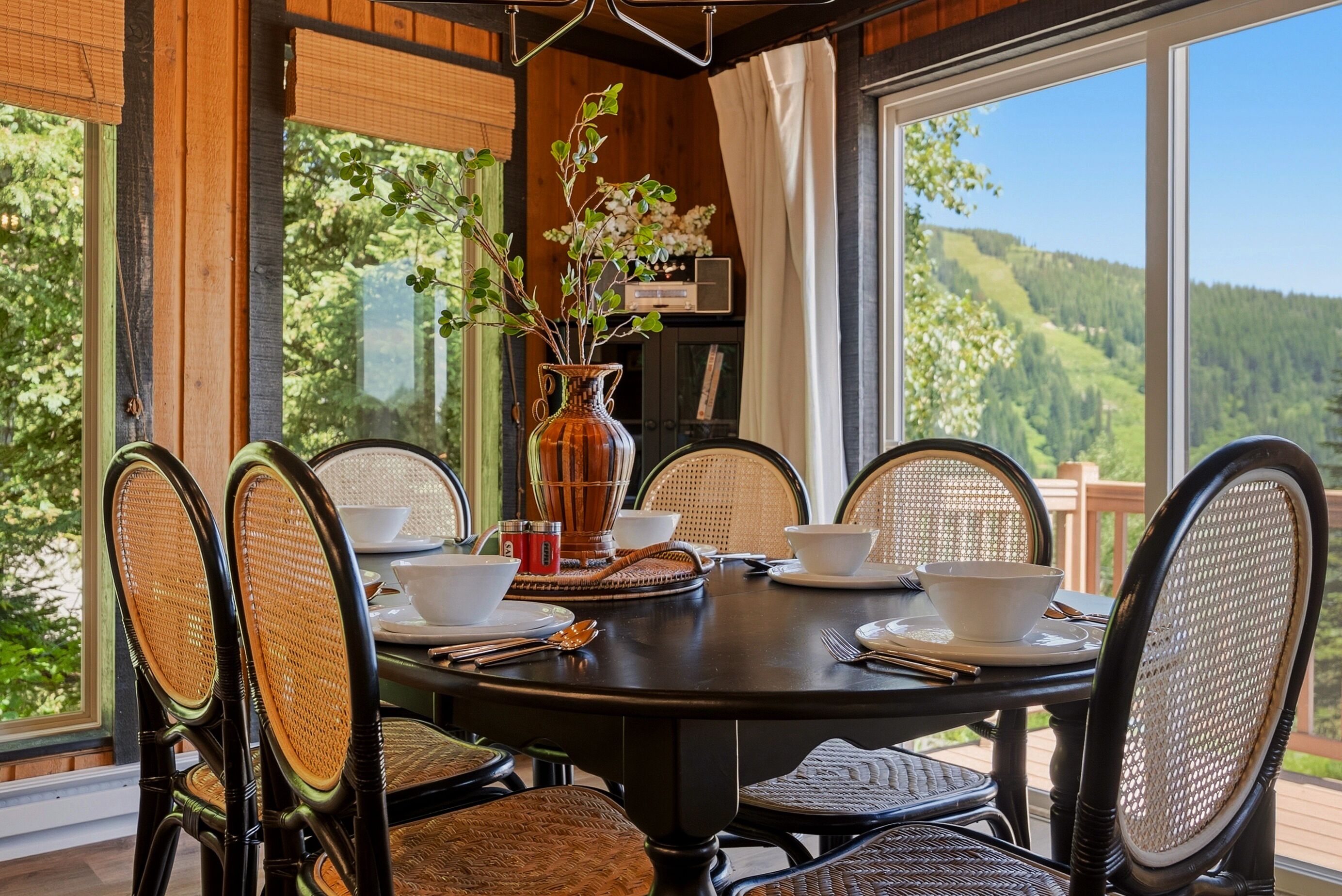 Uma mesa redonda com seis cadeiras de vime, um vaso com galhos e vista para uma paisagem arborizada através de grandes janelas.