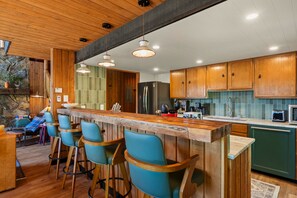 A modern kitchen with wooden cabinets, a bar area with stools, and a stone wall. - Schweitzer Mtn. Retreat -Chalet 75-w/Hot Tub Room + Lift Pass (Sandpoint)