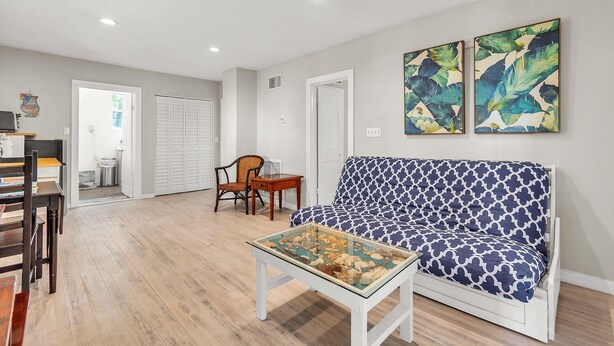 A living room with a sofa, a coffee table with a glass top, a dining area with chairs, and a kitchen visible in the background. - Caddy's Cottage #1 (Treasure Island)