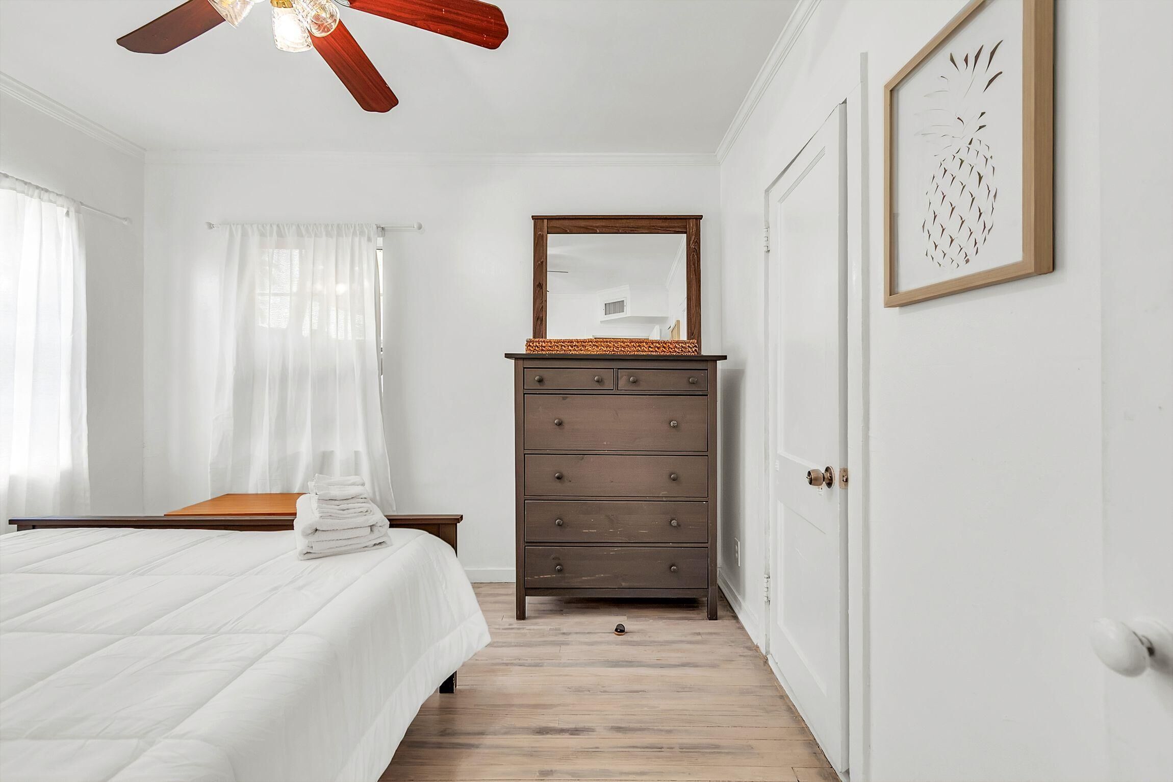 Une chambre à coucher comprenant un lit, une commode, un ventilateur de plafond, un miroir et un tableau représentant un ananas, encadré.