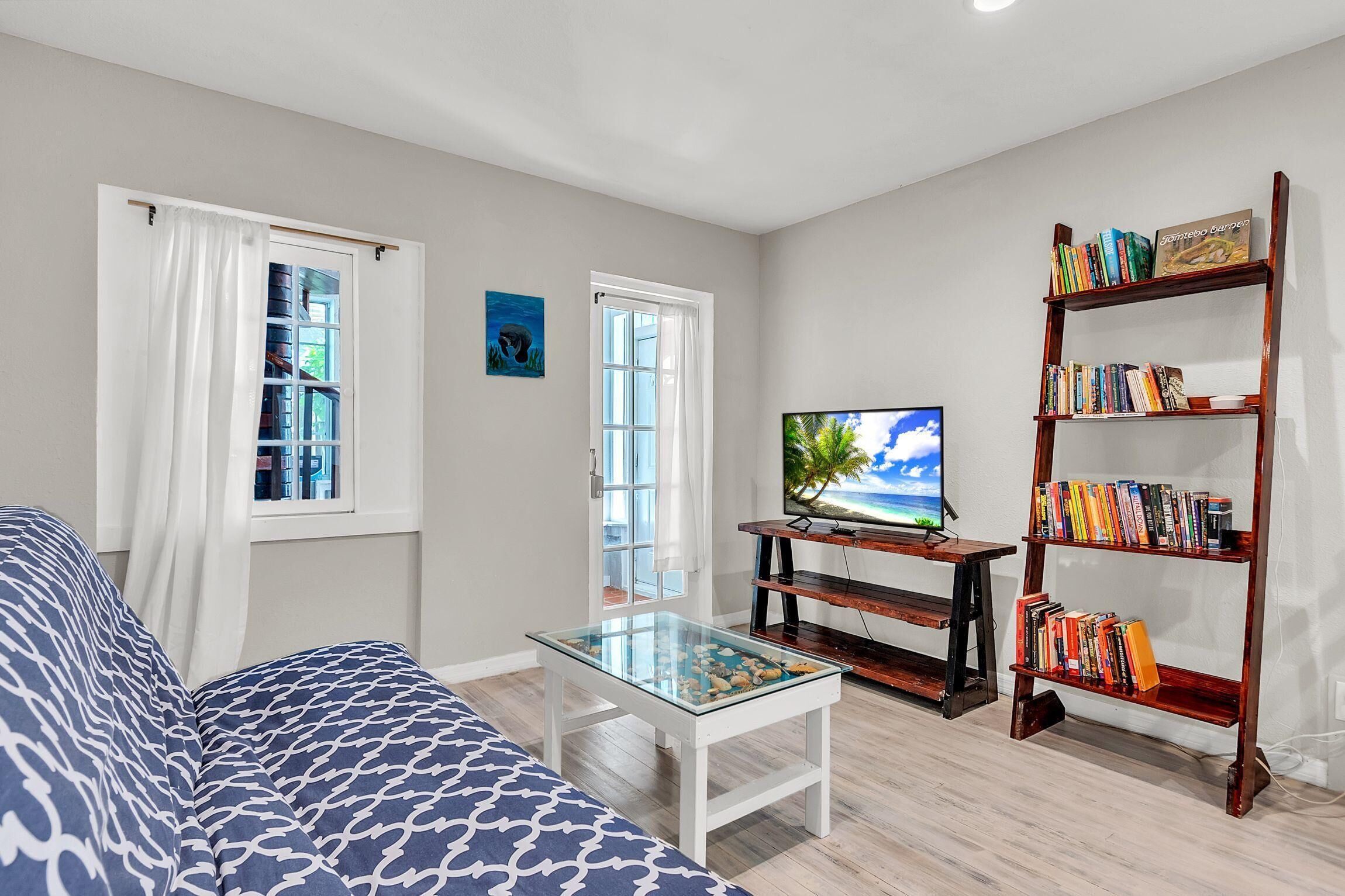Un salon comprenant un canapé, une table basse en verre, une bibliothèque, un téléviseur et deux fenêtres avec des rideaux.