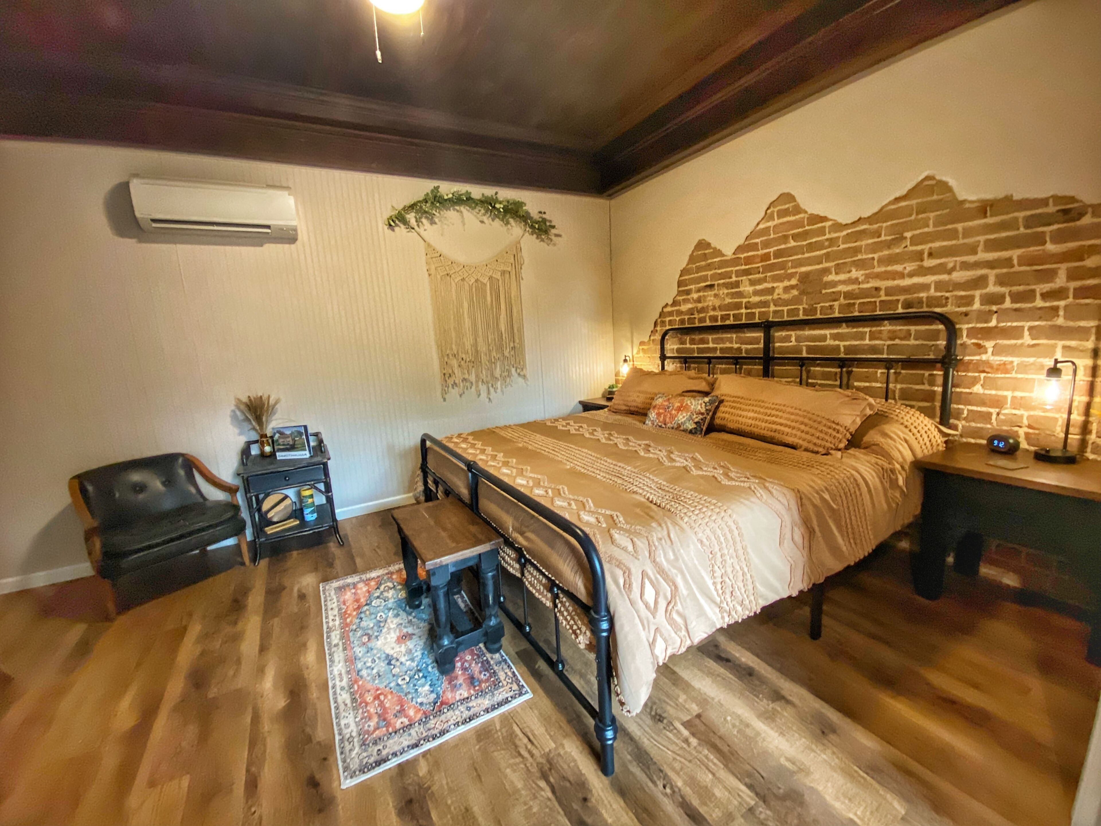 A bedroom with a brick-patterned headboard, a bed with patterned bedding, a small bedside table with a lamp, a chair, and a decorative wall hanging.