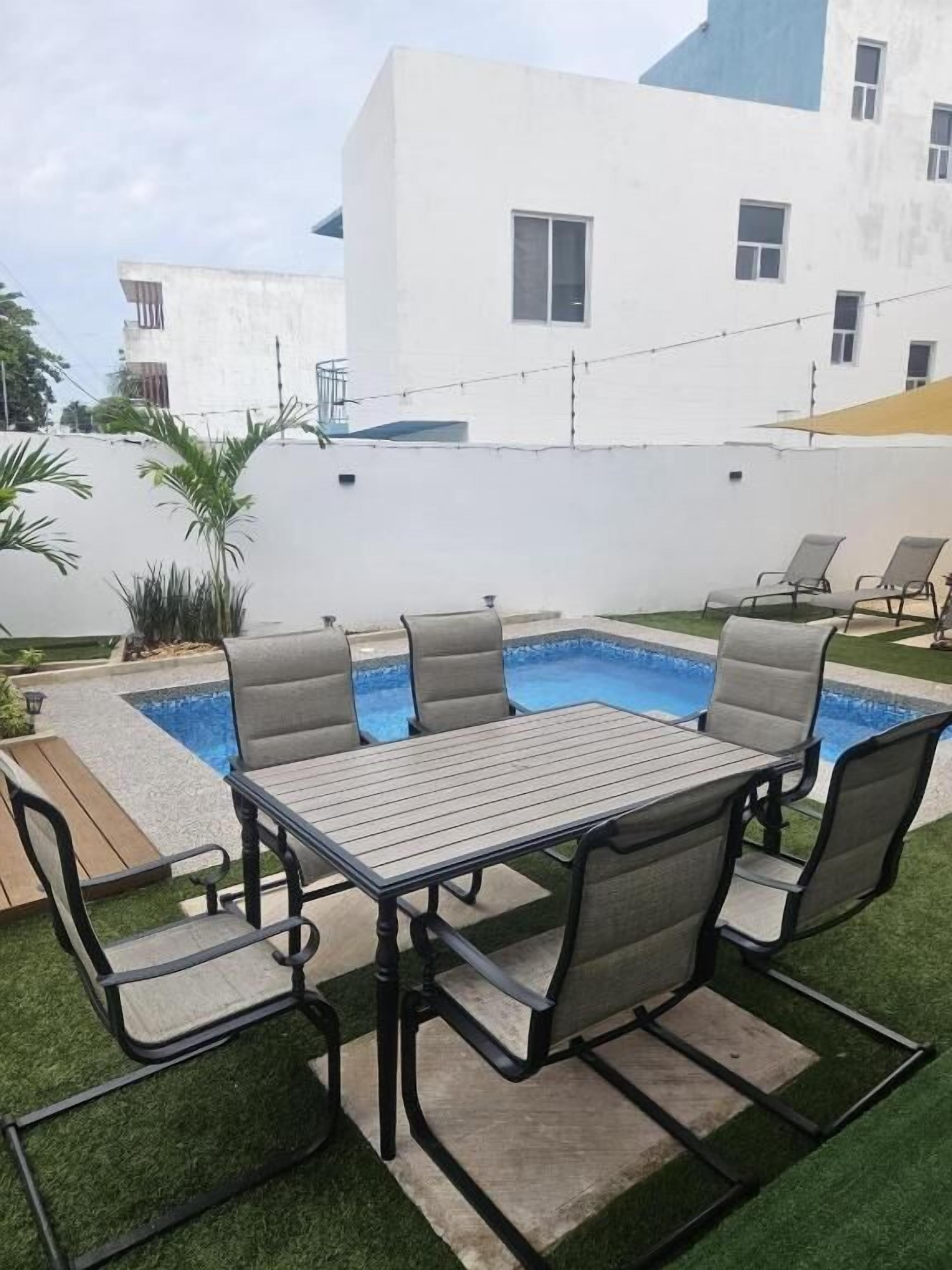 A poolside area with a table and chairs, artificial grass, and a white building in the background.