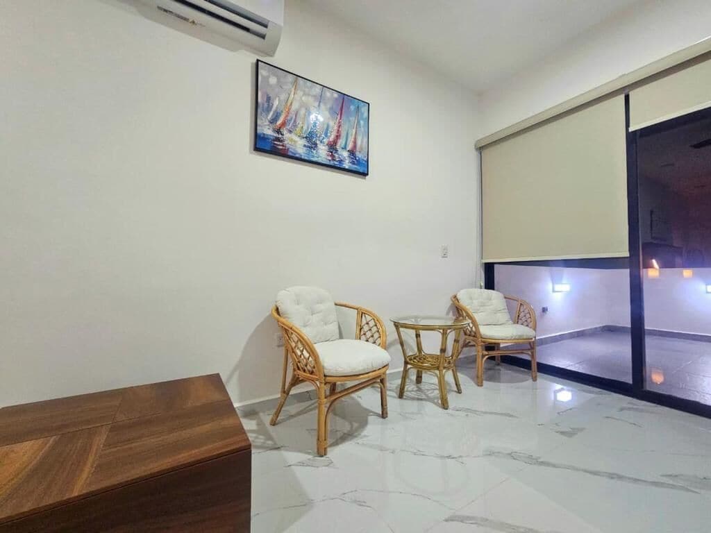 A room with a wooden coffee table, two wicker chairs, a painting of sailboats, and a large window with a view.