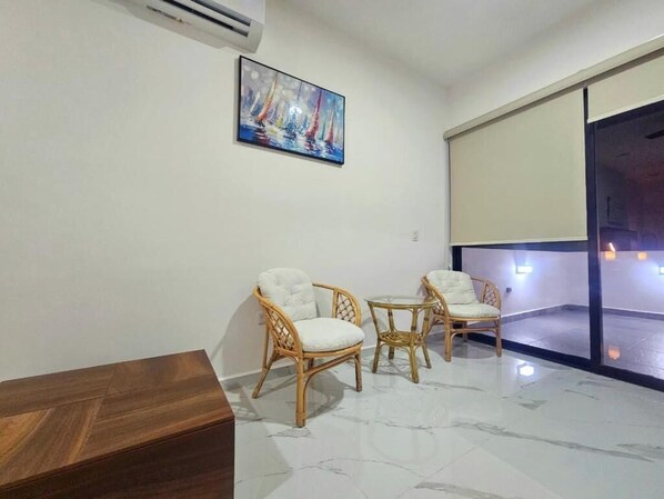 A room with a wooden coffee table, two wicker chairs, a painting of sailboats, and a large window with a view.
