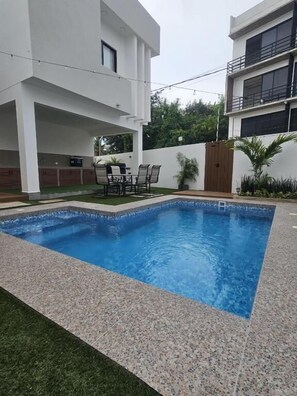 Uma piscina com deck ladrilhado, área de pátio com mesa e cadeiras, e um edifício de vários andares ao fundo.