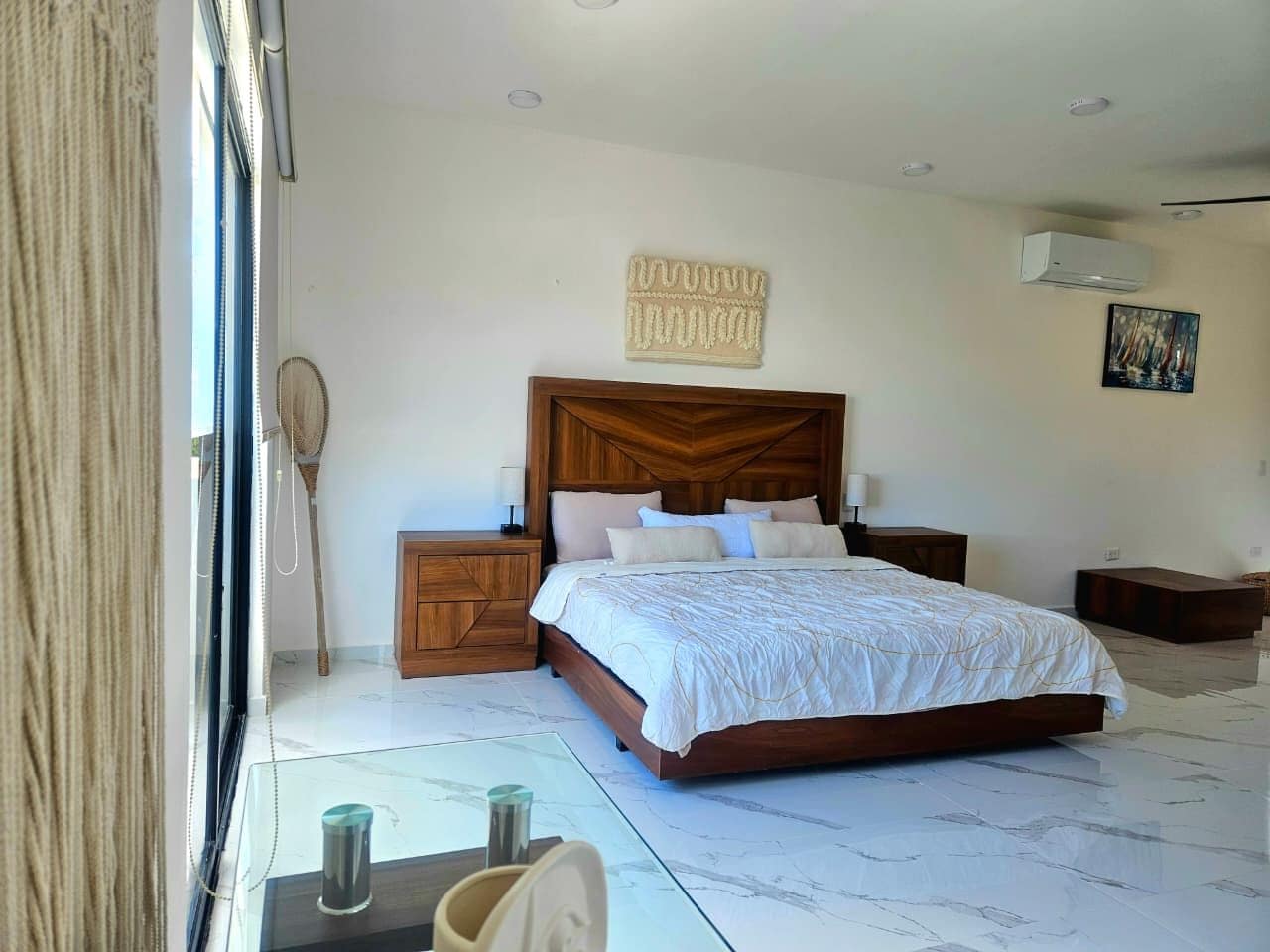 A bedroom with a large bed, wooden headboard, bedside tables, a wall-mounted air conditioner, and a window with curtains.